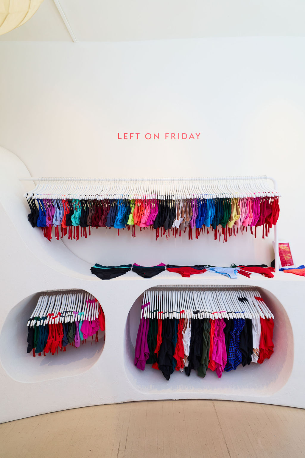 A white retail display wall labeled 'LEFT ON FRIDAY' holding many racks of colorful underwear or bikini bottoms, with several pieces draped on the shelf in front. The setting appears to be a clean boutique interior with light wood flooring.