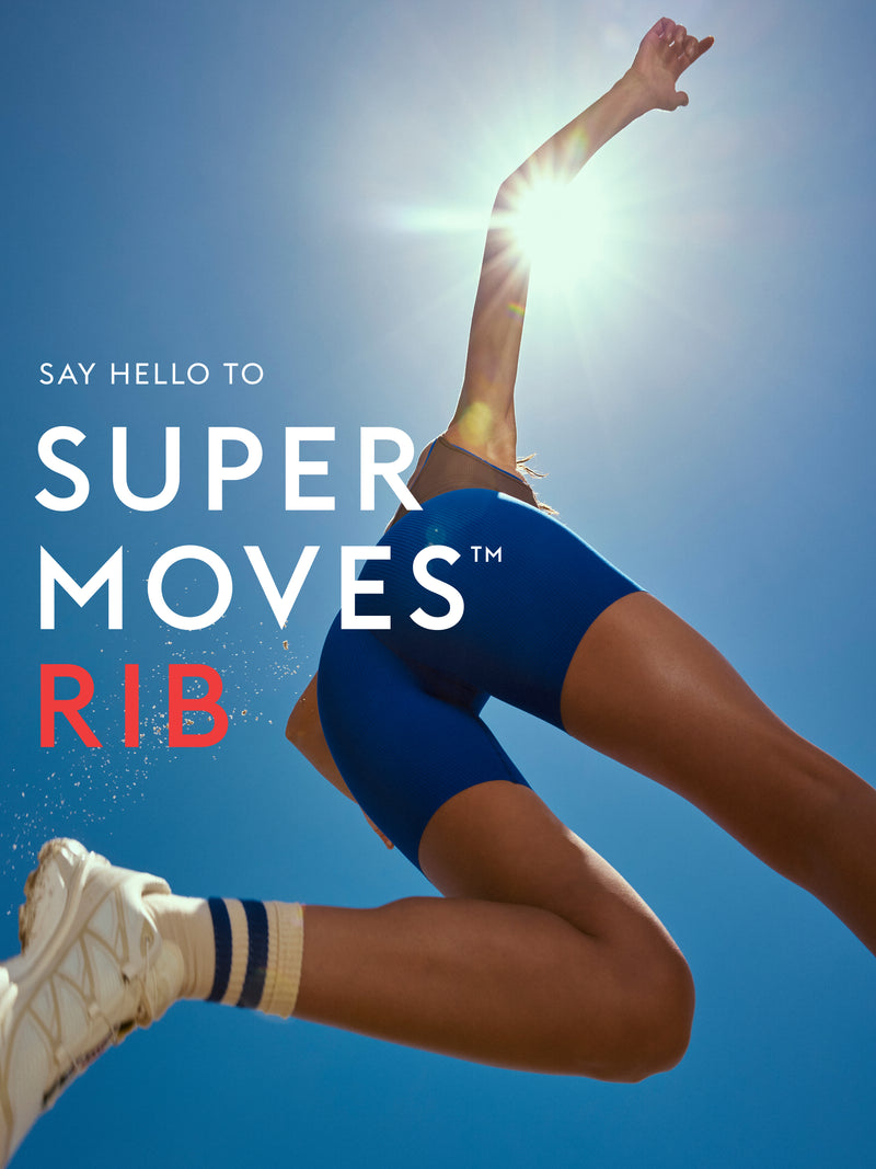 A dynamic lifestyle shot of a female athlete mid-jump against a bright blue sky with the sun shining behind her. She is wearing blue Super Moves Short, white sneakers, and striped socks.