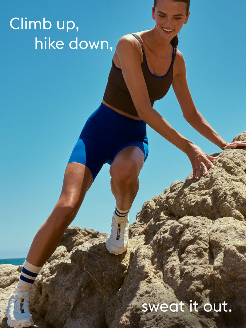 A woman in athletic wear climbing rocks outdoors under a clear blue sky. She is wearing a dark brown Barre Tank, bright blue fitted shorts, white sneakers, and striped socks.