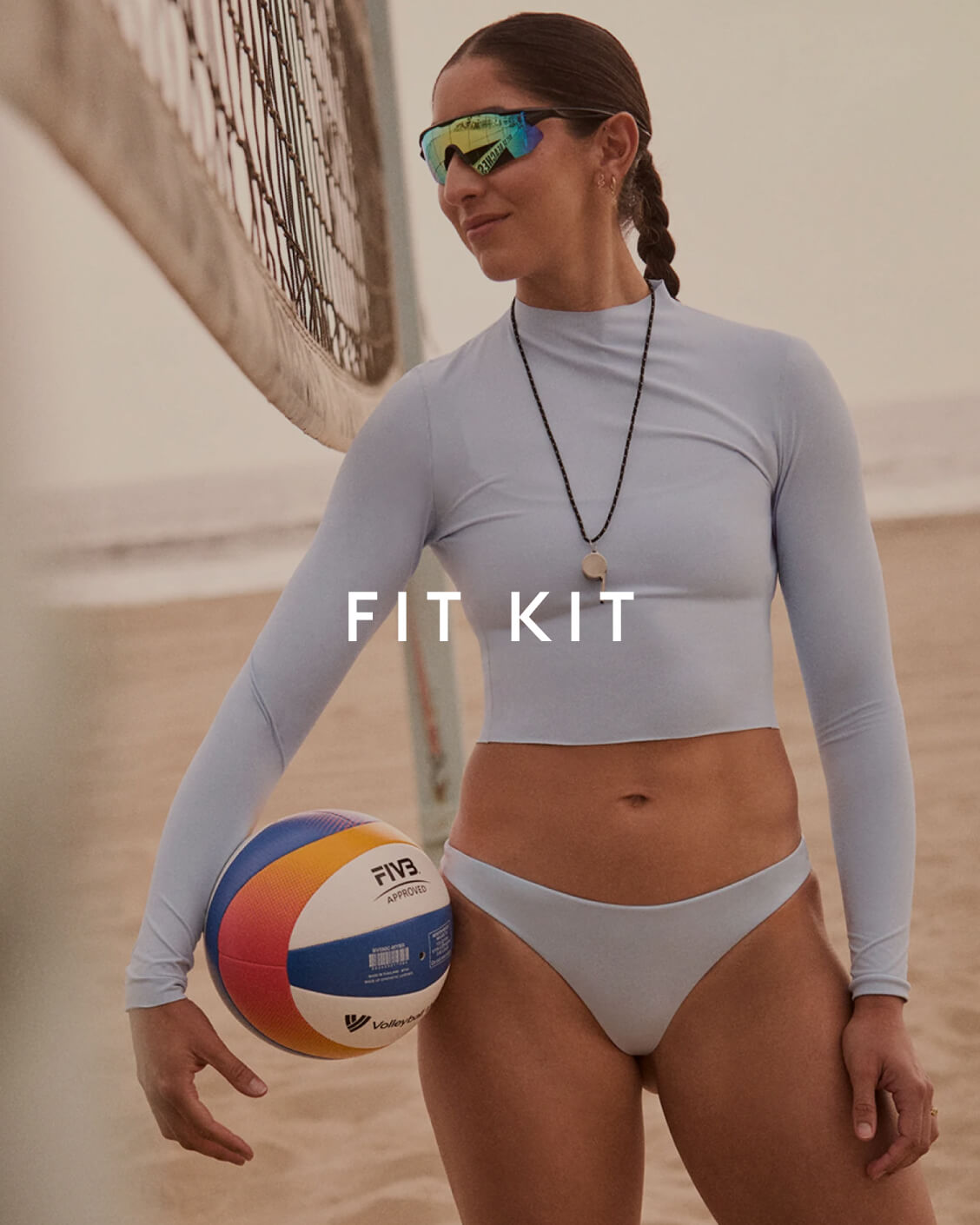 A woman on a beach wearing a light gray long-sleeve crop top and matching bikini bottoms, holding a FIVB volleyball. The text 'FIT KIT' is overlaid on the center of the image.