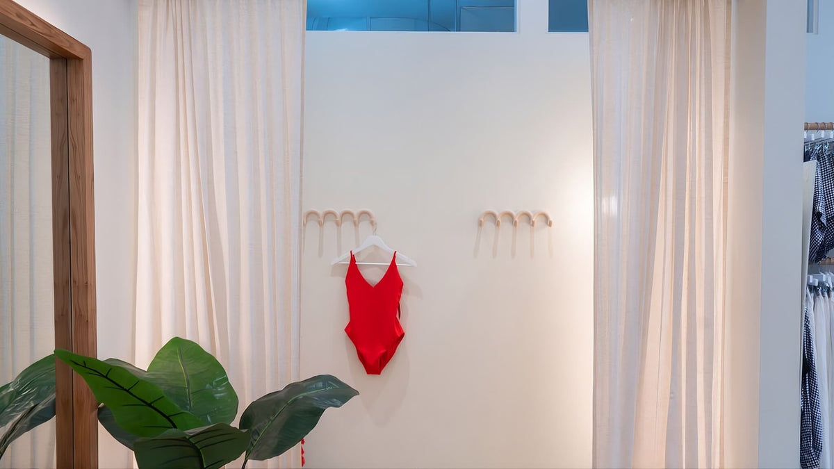 A bright red one-piece swimsuit hangs on a white wall in a fitting room with white curtains, a wooden mirror frame on the left, and a green plant in the foreground.