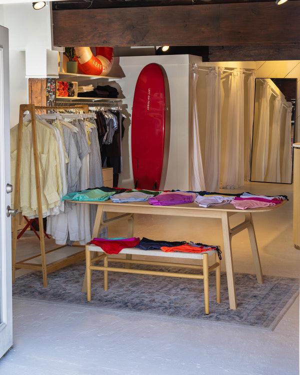 A retail clothing store interior with a wooden table displaying folded colorful garments, a matching wooden bench with folded clothes on it, hanging shirts on racks, a red surfboard leaning against a wall, and beige curtains in the background.