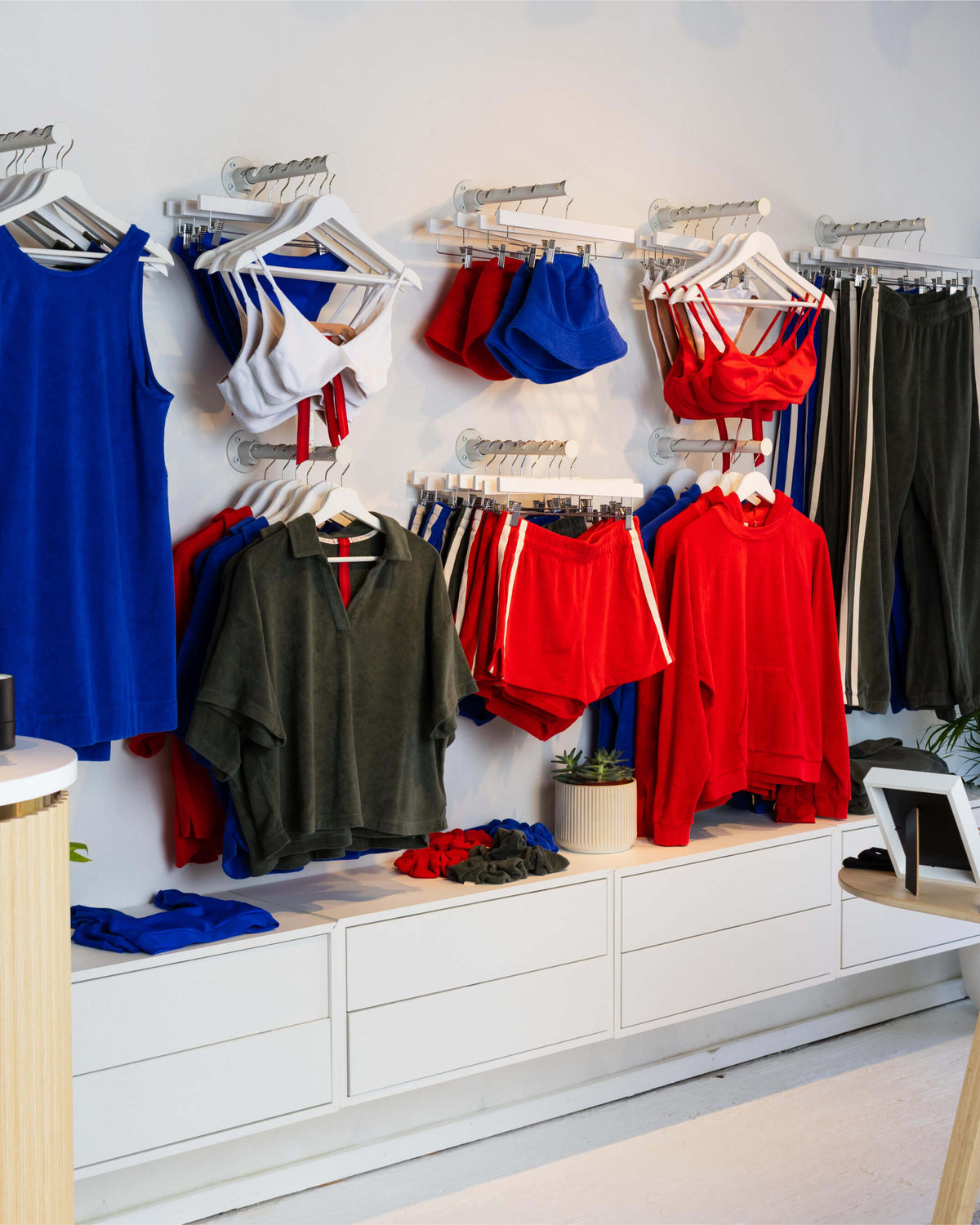 A retail clothing display featuring multiple garments on white hangers mounted on a wall above white storage drawers. Visible items include blue, red, white, and dark green tops, shorts, and pants.