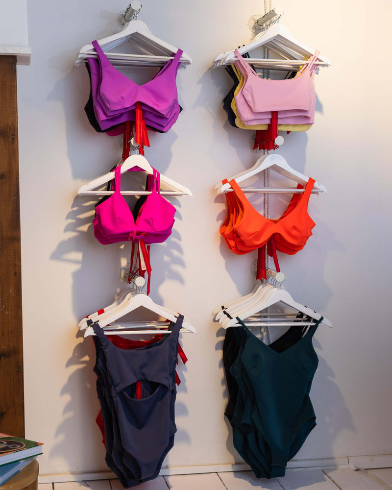 Six swimsuits in various colors (purple, pink, orange, navy, and teal) are displayed on white hangers hanging from a white wall.