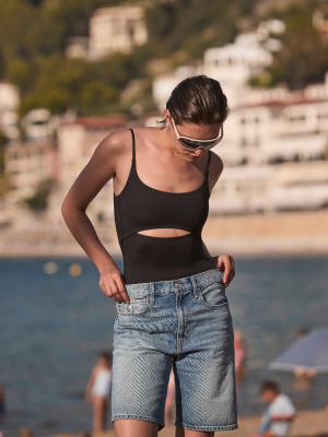 Person wearing sunglasses and a black Peak Suit with denim shorts by a waterfront.