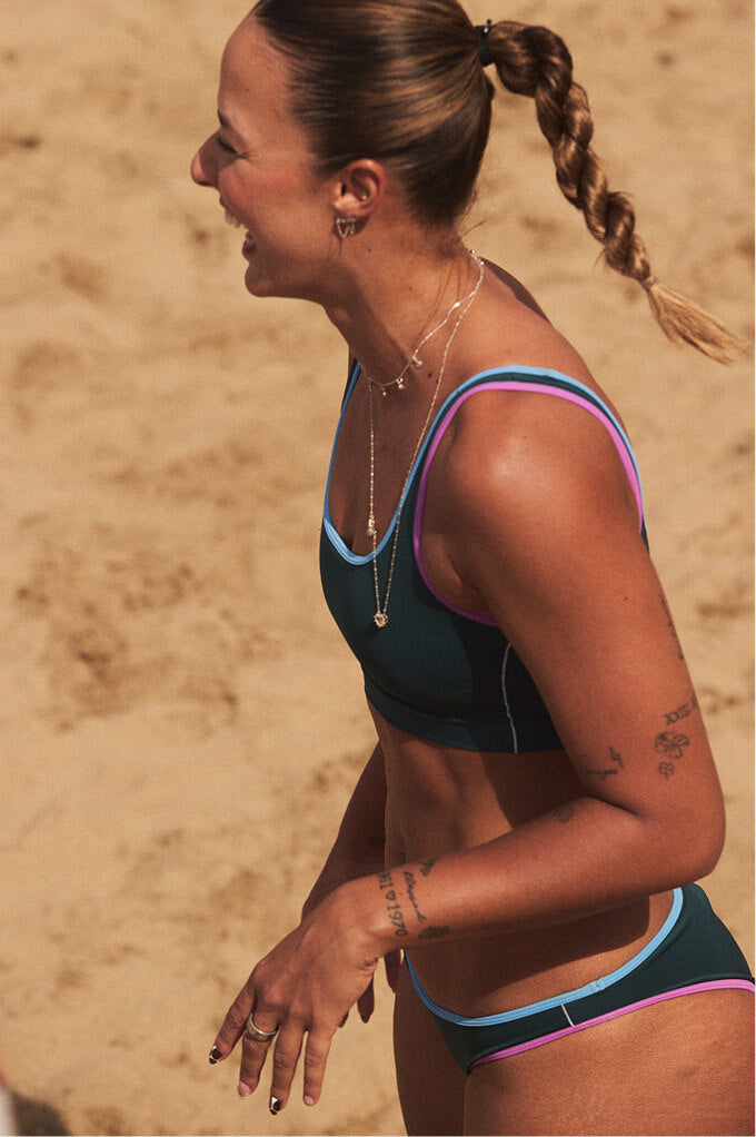 A woman in a dark teal bikini with pink and light blue trim stands on a sandy beach, shown in profile with her hair in a braid.