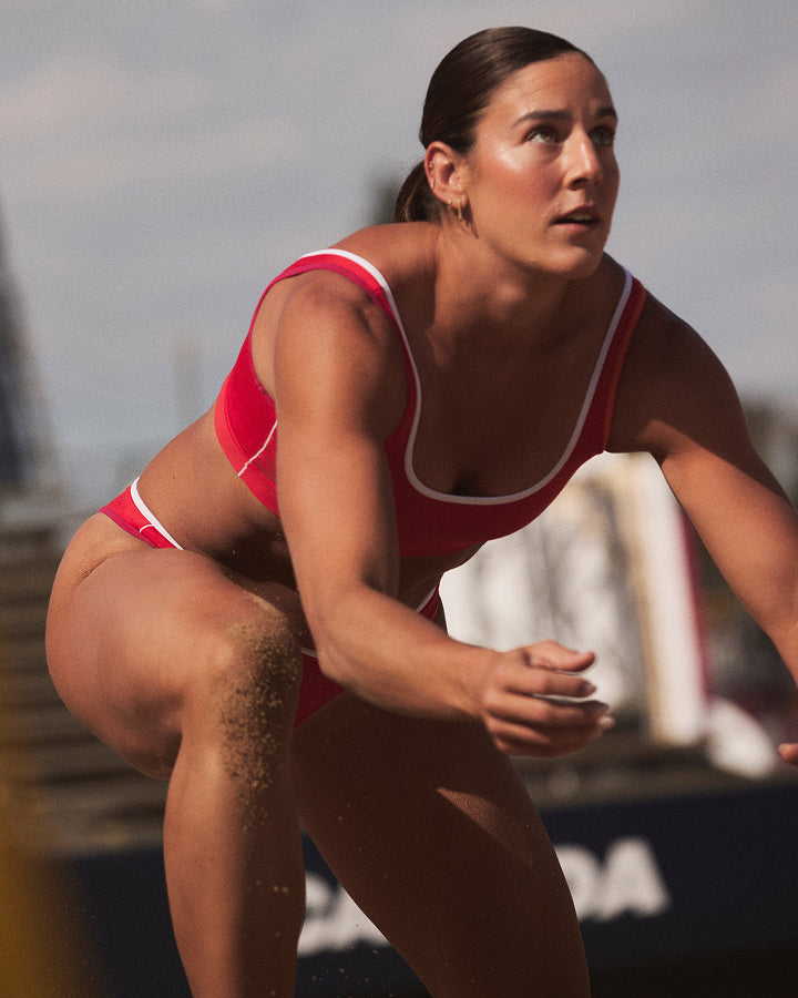 A woman in a red and white bikini swimsuit crouches in a water sport setting, with sand visible on her leg and a blurred outdoor background.