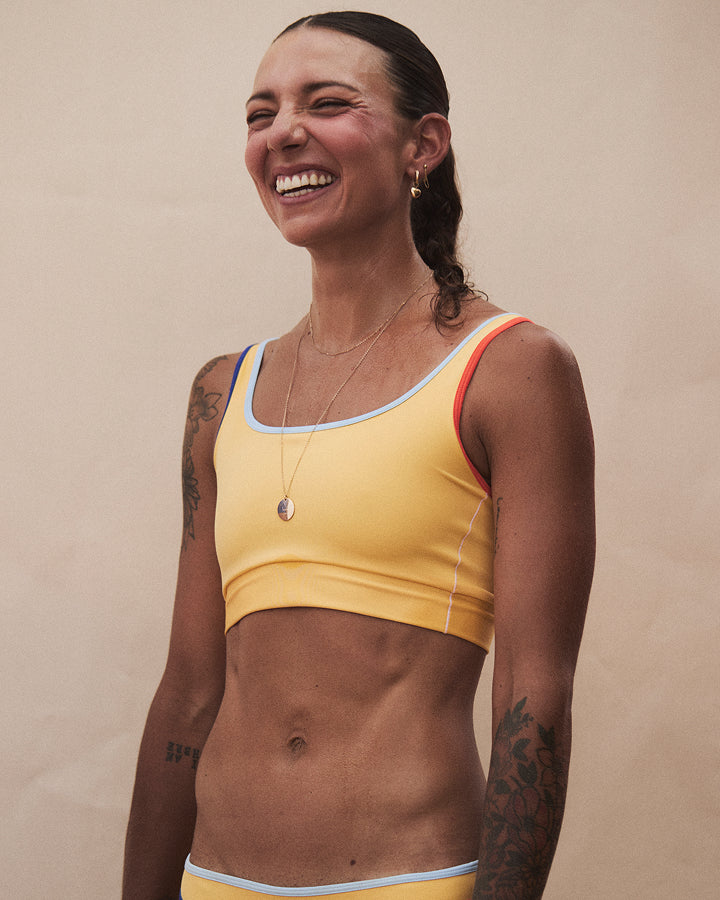 A woman smiling and looking to the side, wearing a yellow athletic sports bra with light blue and orange trim, set against a plain beige background.