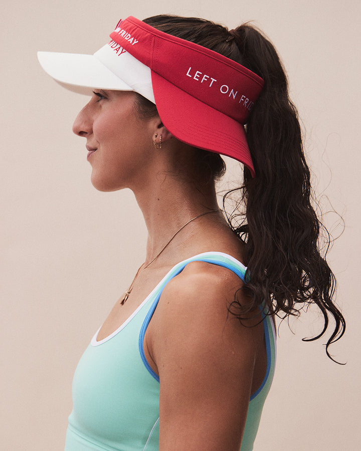 A woman in profile wearing a red and white visor with the text 'LEFT ON FIRE' and 'EVERYDAY' on the brim, paired with a light blue and white sports bra, against a solid beige background.