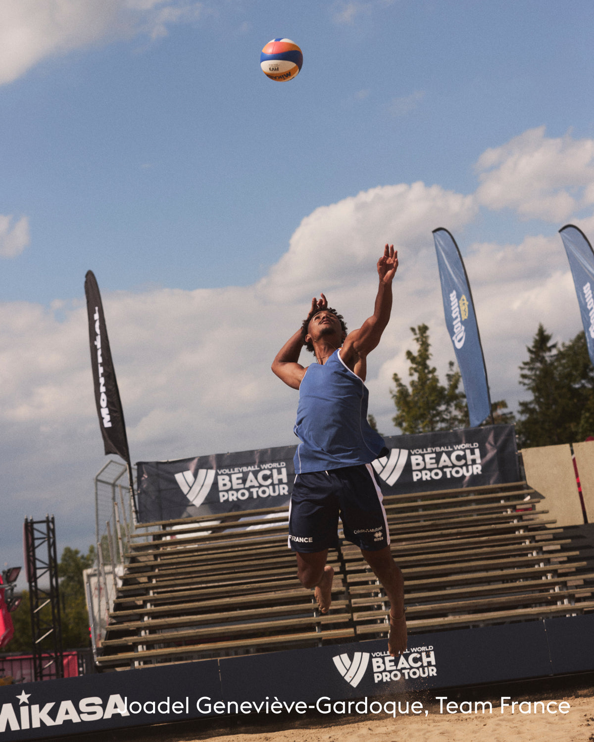 A beach volleyball player in a blue sleeveless top and dark shorts jumps to spike a ball over a sand court, with bleachers, banners, and a blue sky in the background.