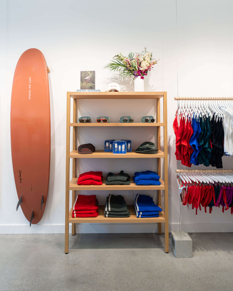 A wooden four-tier shelf in a retail store setting, displaying folded colorful clothing, small accessories, and a vase of flowers. A large orange surfboard leans against the wall on the left, and a clothing rack with red, blue, green, and white garments is on the right.