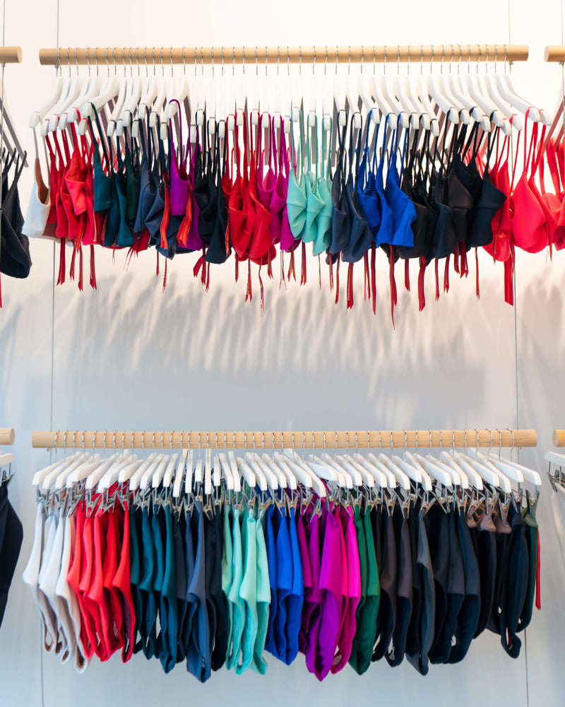 Two wooden clothing racks filled with many colorful bikini or bralette tops hanging on white hangers against a white wall.