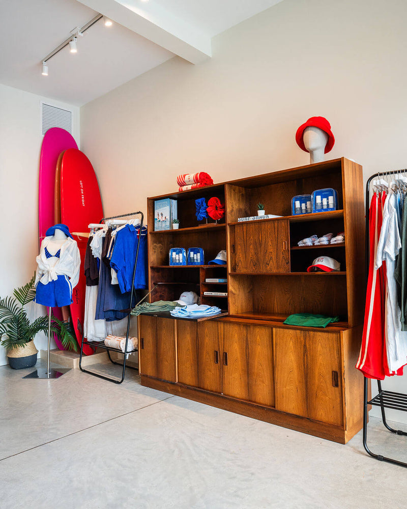 A wooden display cabinet in a retail store, filled with folded and hanging towels, hats, and small blue boxes, alongside clothing racks and bright surfboards against a beige wall.