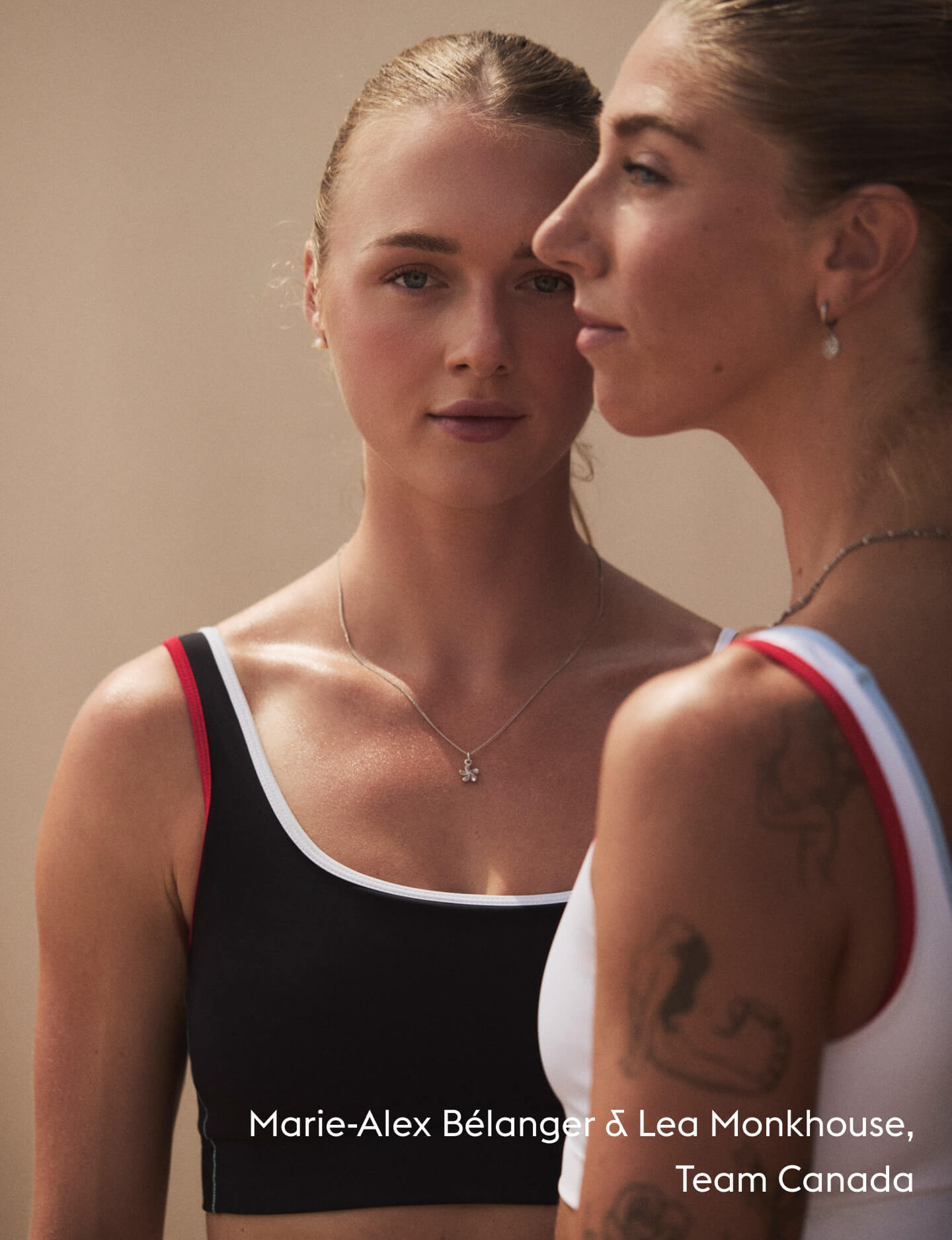 Two female athletes in athletic wear, one wearing a black sports bra with white and red trim, and the other wearing a white sports bra with red and black trim, set against a plain beige background.