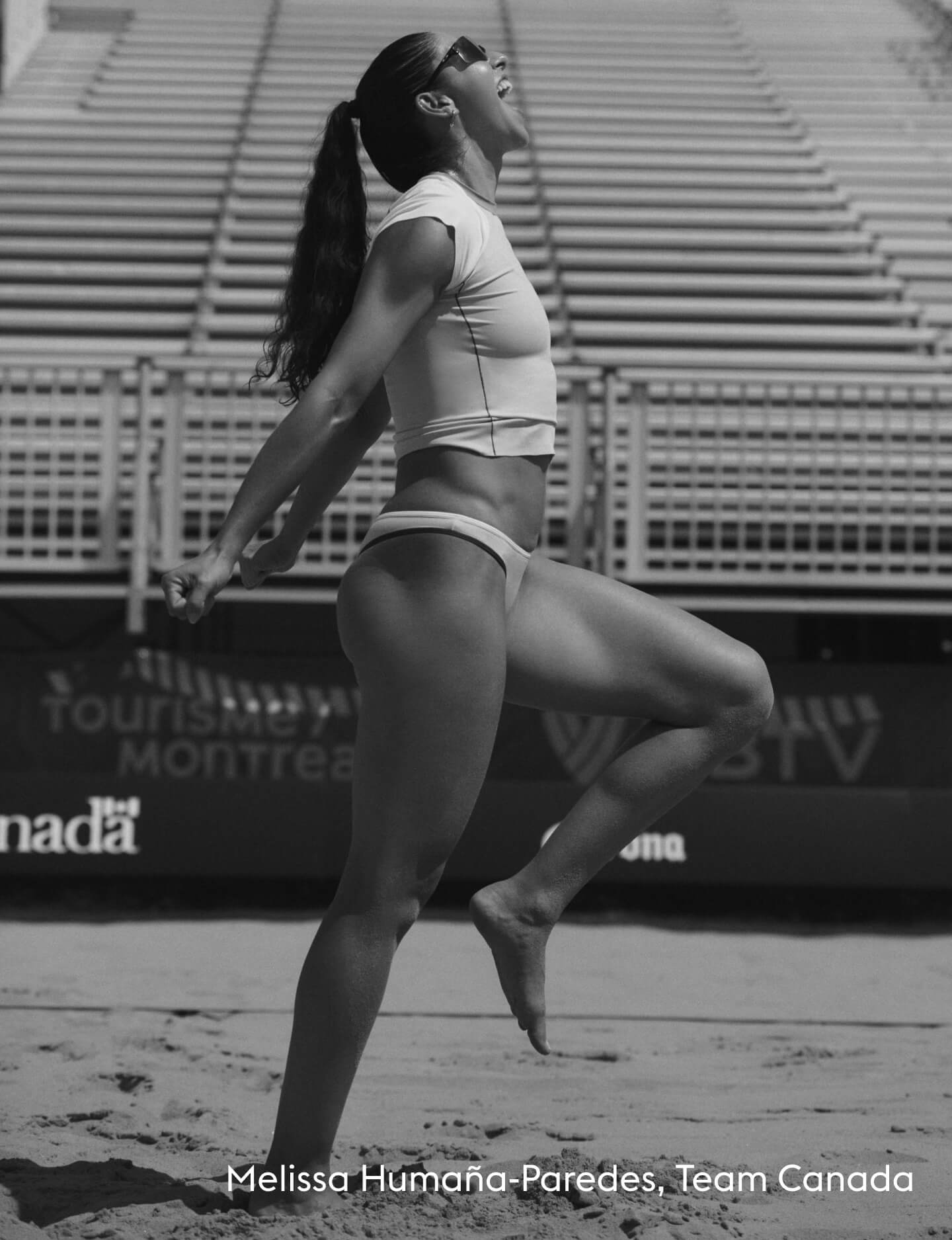 Black and white photo of Melissa Humana-Paredes in a white bikini posing on a sand court with stadium seating in the background. Text at the bottom reads 'Melissa Humana-Paredes, Team Canada'.
