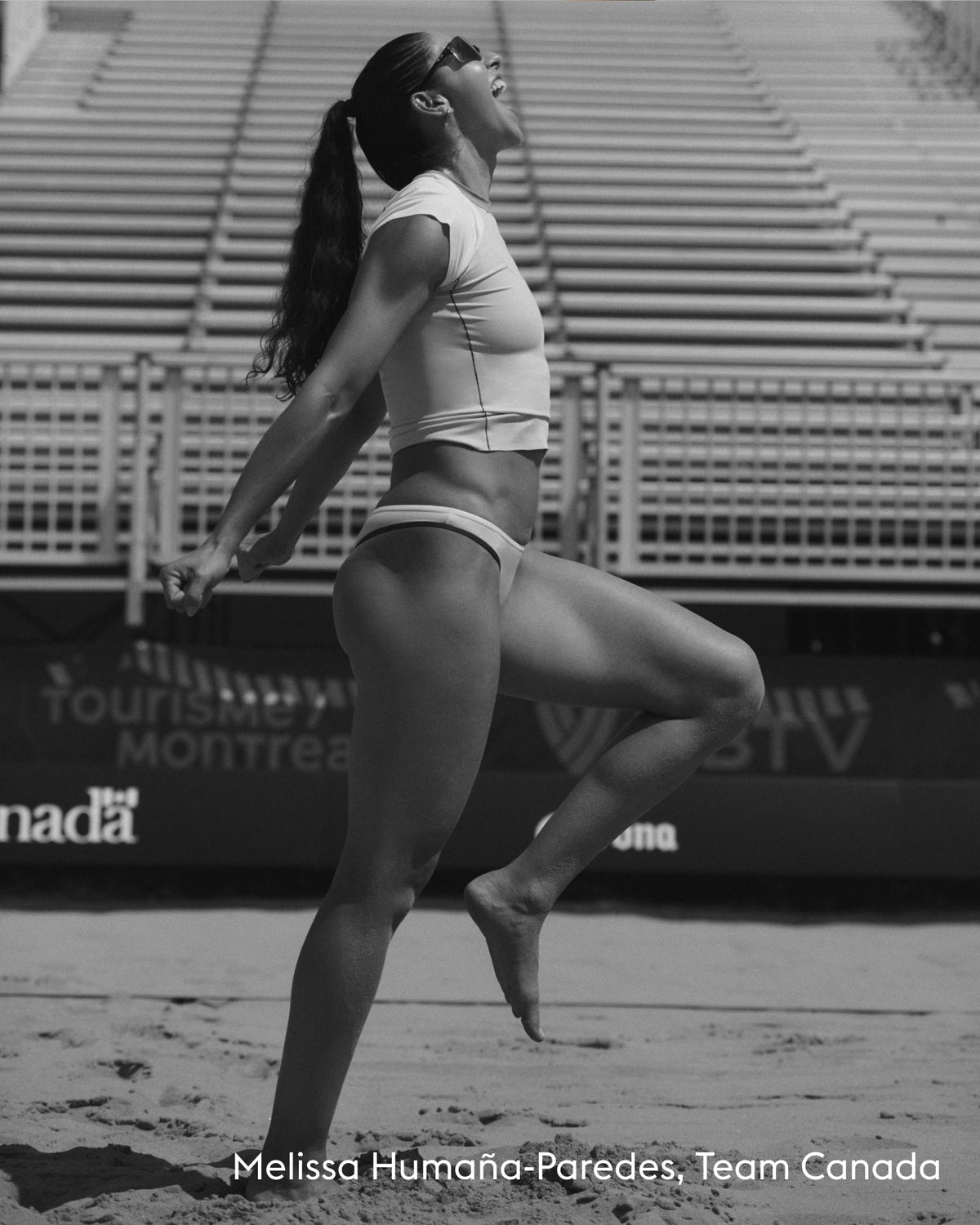 Black and white photo of Melissa Humana-Paredes in a white crop top and bikini bottoms, captured mid-jump on a beach court with stadium seating in the background. Text at the bottom reads 'Melissa Humana-Paredes, Team Canada'.