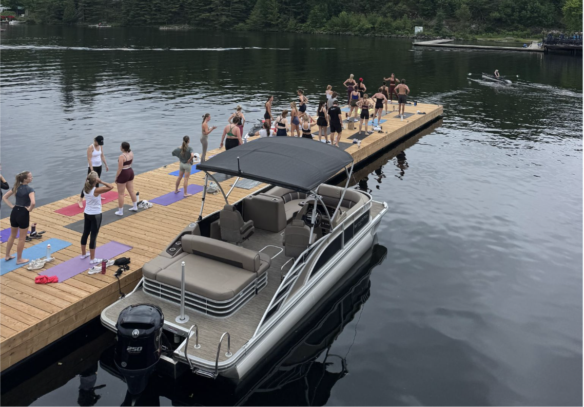 A silver and gray pontoon boat with a black canopy is docked at a wooden pier where a group of people are practicing yoga on colorful mats. The scene is set on a lake with trees in the background.