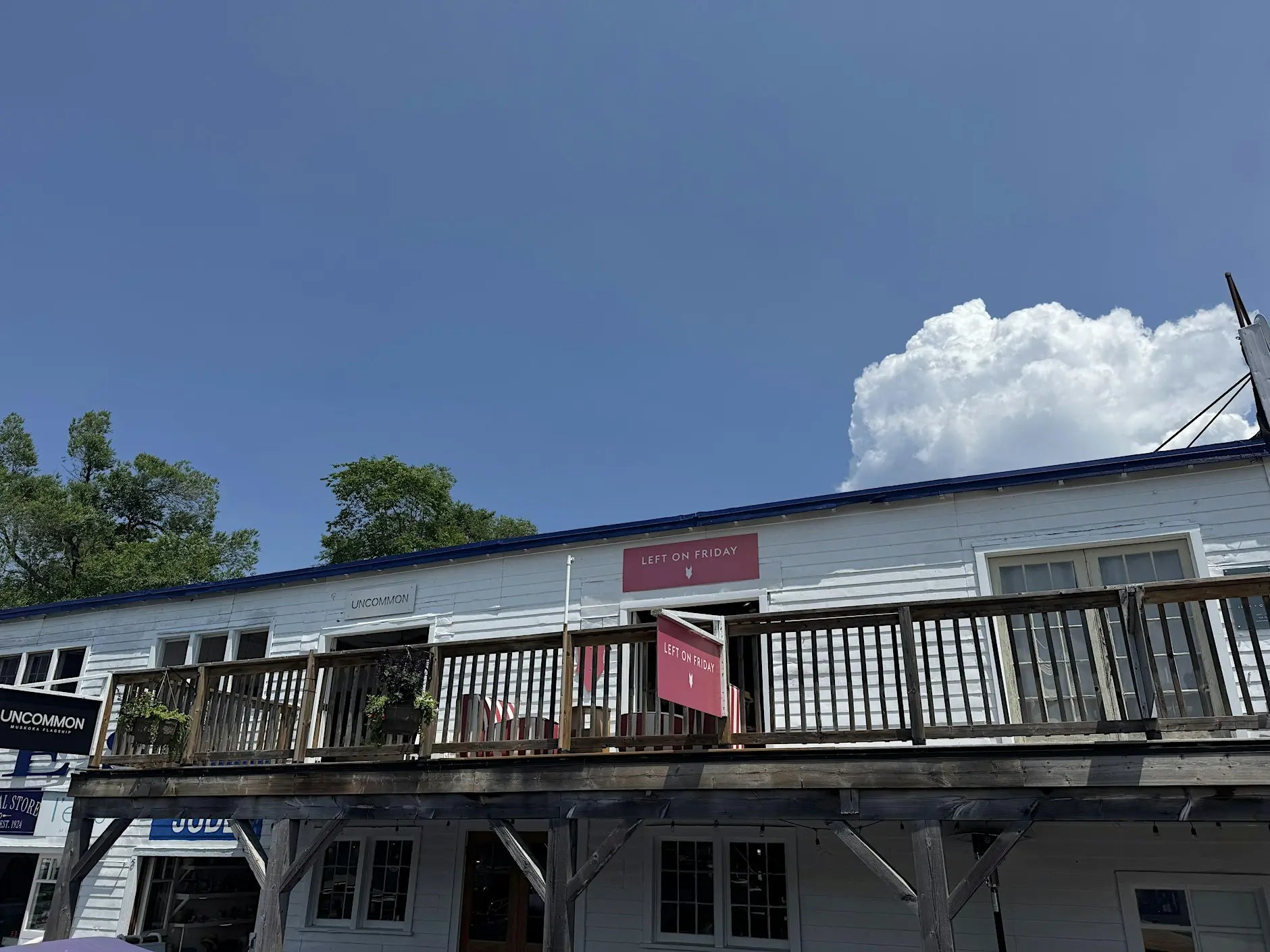 A white wooden building with a dark blue roof and a raised wooden balcony, photographed outdoors under a bright blue sky with a large white cloud. A red sign is visible above the entrance.